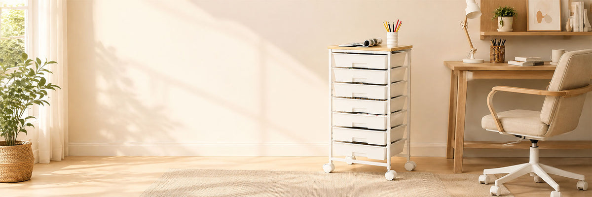 A white rolling storage organizer with multiple drawers stands in a bright, minimalist home office next to a wooden desk and beige office chair.