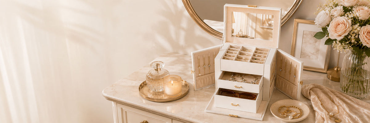 A cream-colored wooden jewelry organizer with multiple drawers and compartments, shown open on a vanity table next to a mirror, flowers, and decorative accessories.